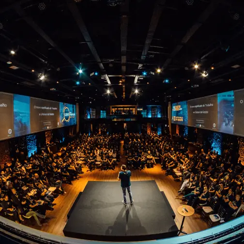 Ein Mann steht auf einer Bühne und spricht zu einem großen Publikum in einem dunklen Auditorium, in dem auf mehreren Bildschirmen Präsentationsfolien angezeigt werden. Durch das Live-Streaming dieses Hybrid Events bietet er sowohl den Teilnehmern vor Ort als auch den Teilnehmern an den entfernten Standorten ein spannendes Erlebnis.