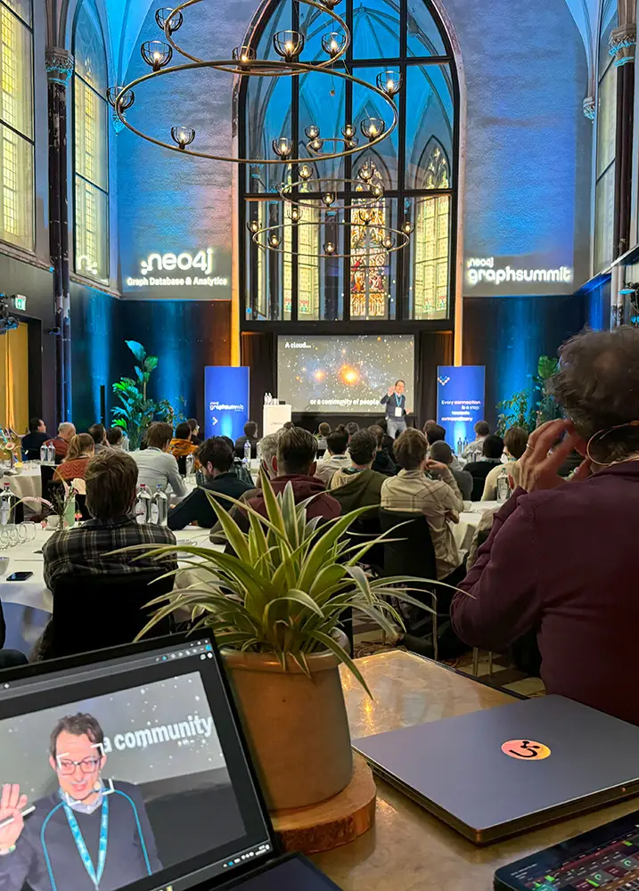 In einer Kirche findet eine Konferenz statt. Die Teilnehmer sitzen vor einer Bühne mit einem Präsentationsbildschirm. Auf einem Tisch im Vordergrund ist ein Laptop zu sehen, auf dem ein Sprecher abgebildet ist. Dies unterstreicht den Aufbau eines Hybrid Events, zu der auch Live-Streaming für Remote-Teilnehmer gehört.