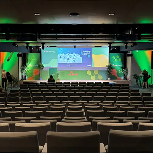 Konferenztechnik mit großer Projektion und grün-oranger Bühnenbeleuchtung im Veranstaltungssaal