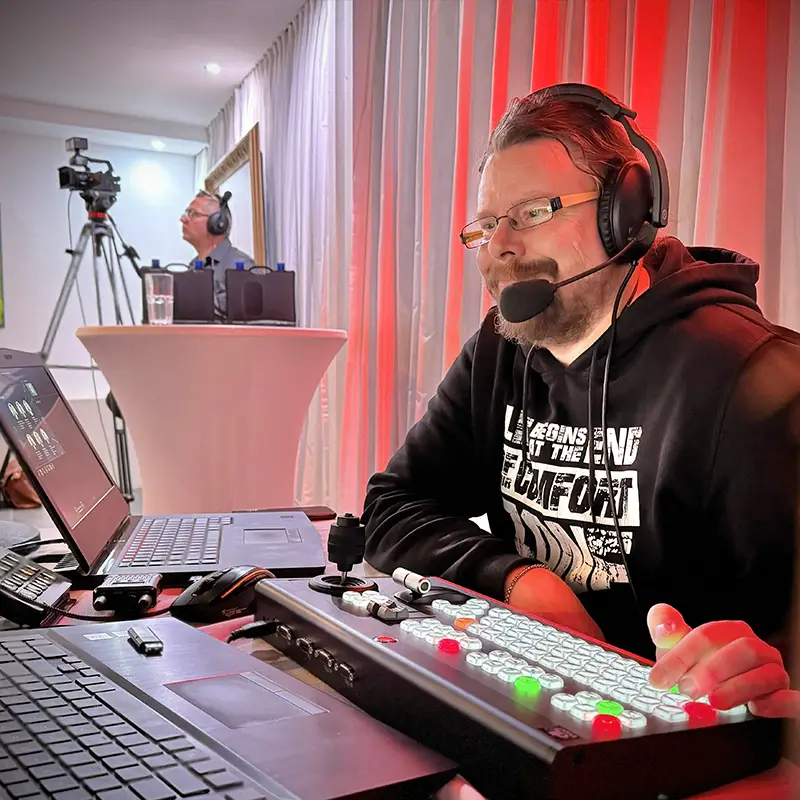 Techniker mit Headset am Bildmischpult einer Livestream-Produktion, im Hintergrund ein weiterer Techniker an einer Stativkamera.