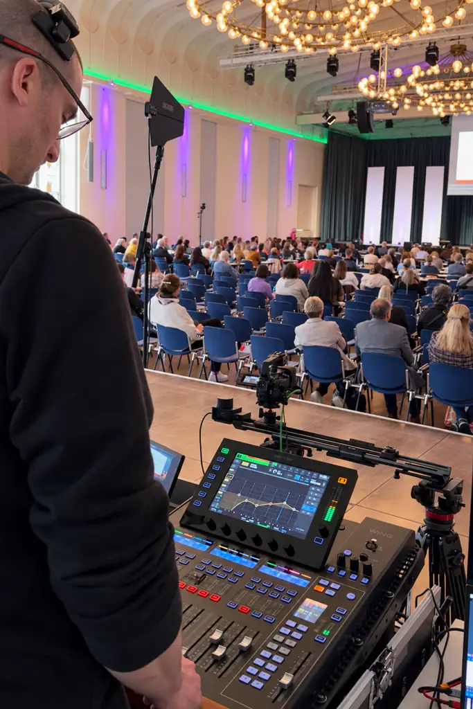 Veranstaltungstechniker am Tonmischpult mit Kamera auf einem Slider und Blick auf den gefüllten Konferenzsaal.