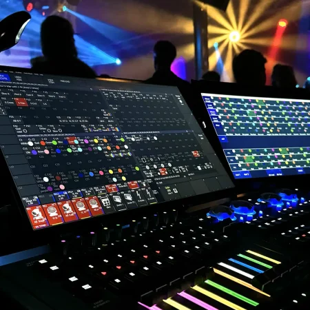 Lichtkonsole mit zwei Bildschirmen und beleuchteten Reglern, dahinter Publikum und farbige Lichtstrahlen einer Liveveranstaltung.